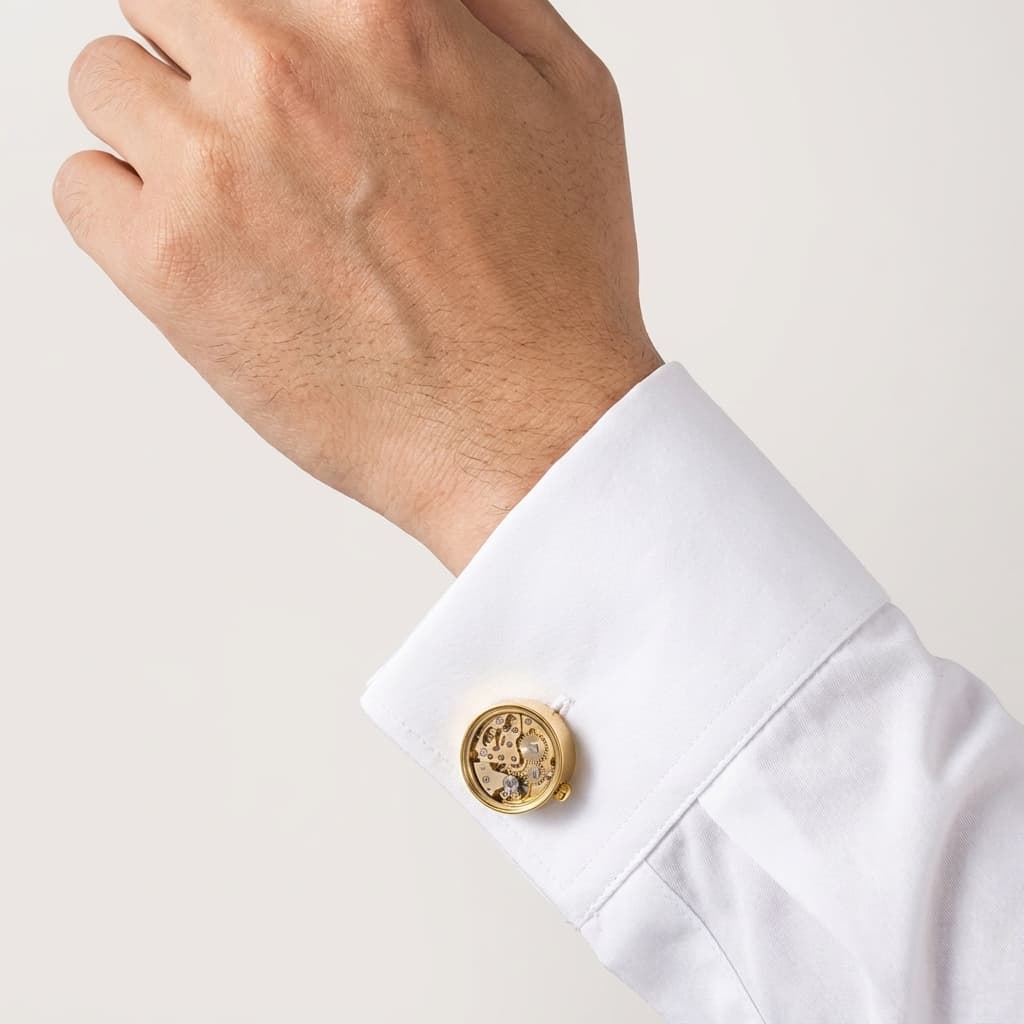 Gold skeleton movement cufflinks with visible brass gears and ruby bearings on a white dress shirt cuff.