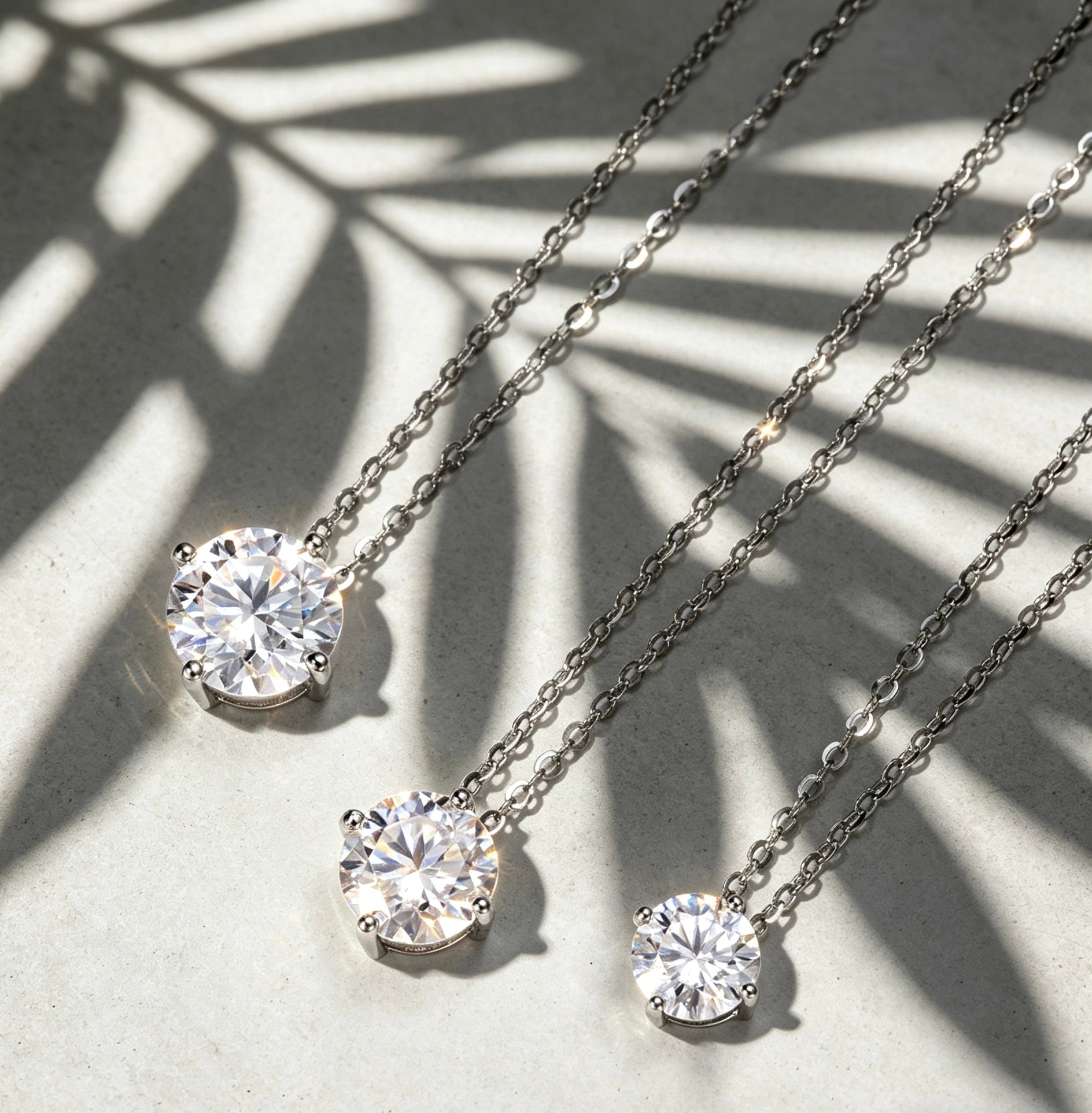 Three sparkling moissanite solitaire necklaces in four-prong S925 silver settings on a textured surface with palm shadows.