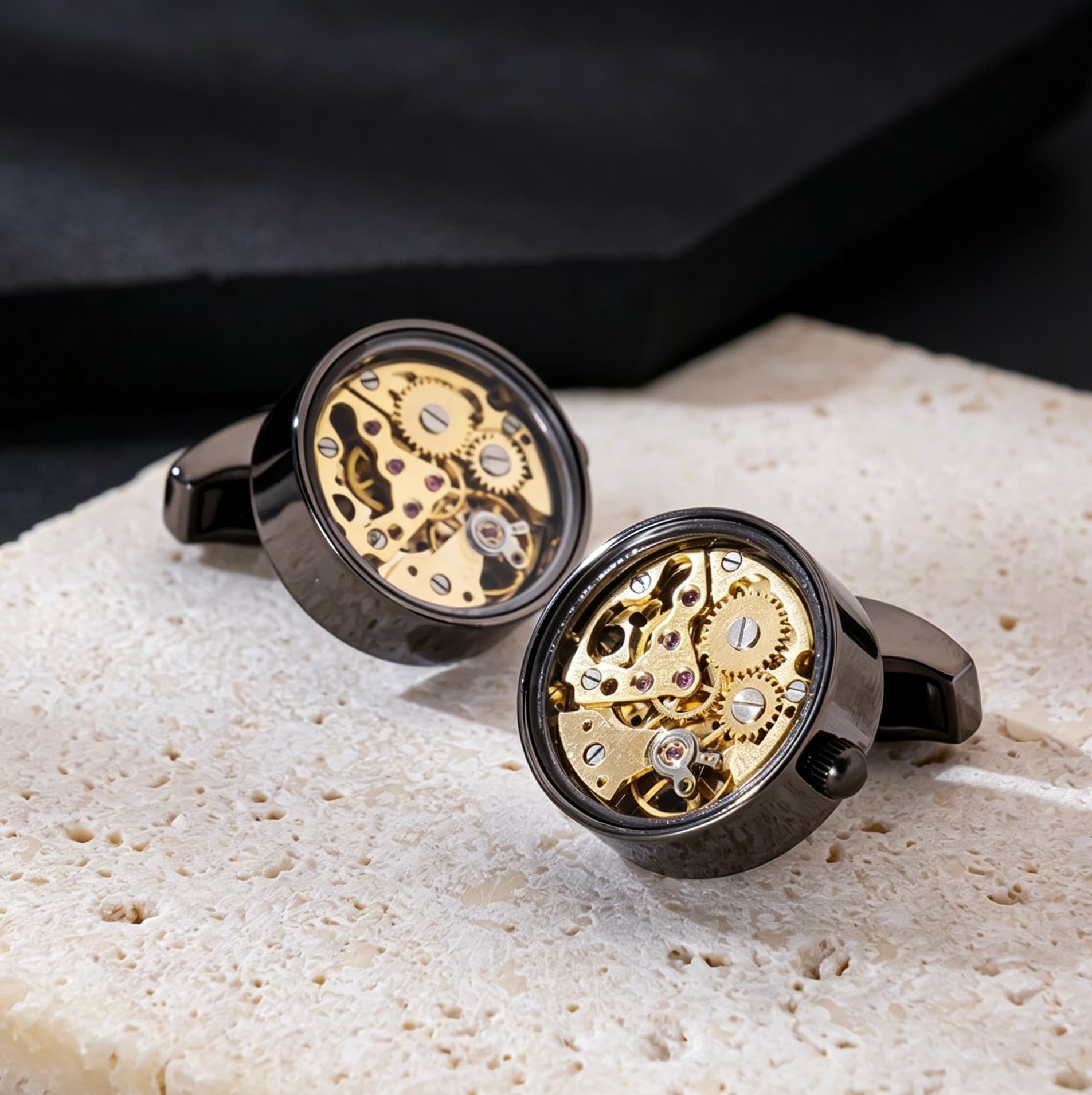 Round brass skeleton movement cufflinks featuring visible gold gears and ruby-red jewel bearings on a textured stone surface.