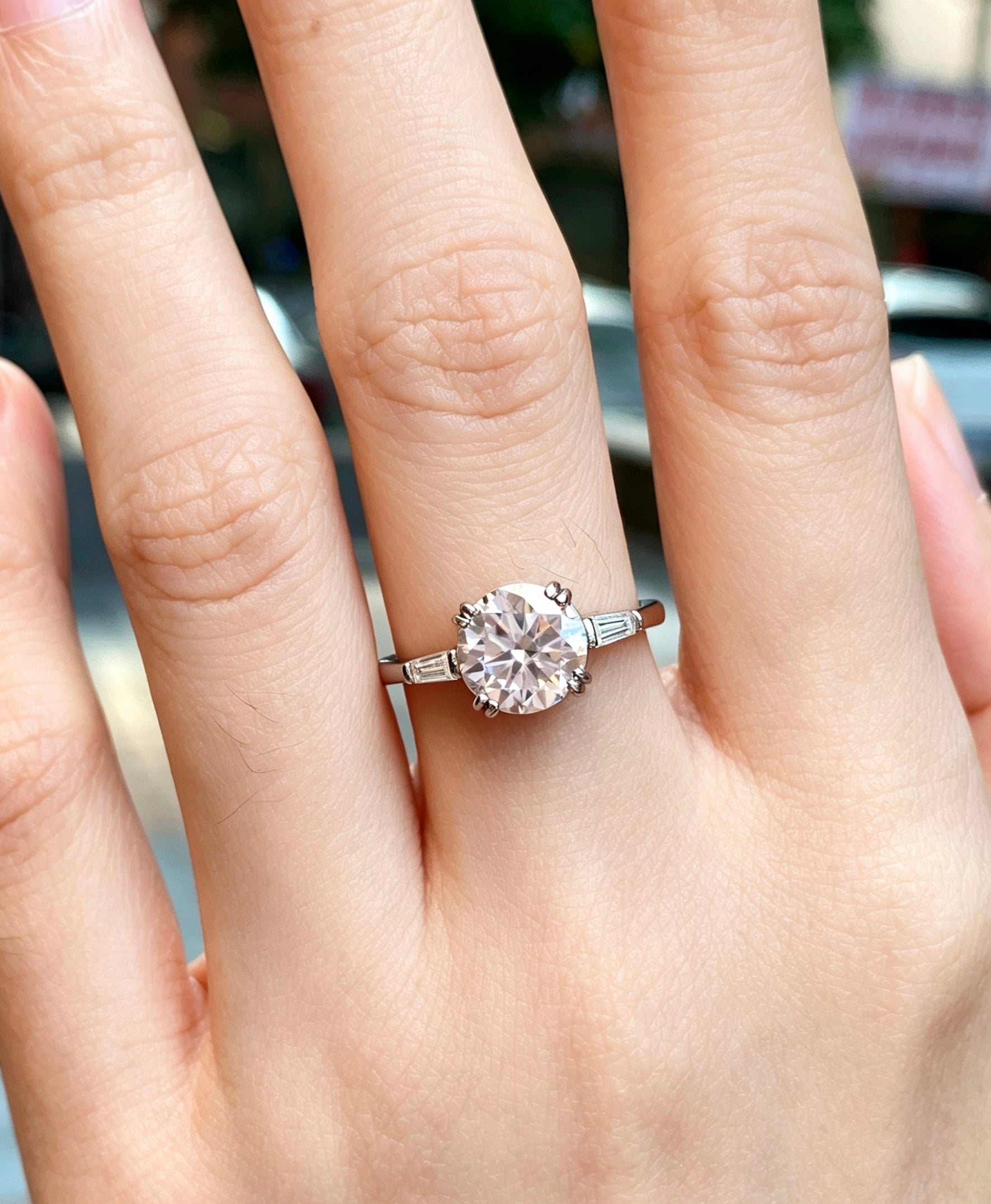 Six-prong round moissanite engagement ring with tapered baguette accents on a silver S925 band worn on a finger.