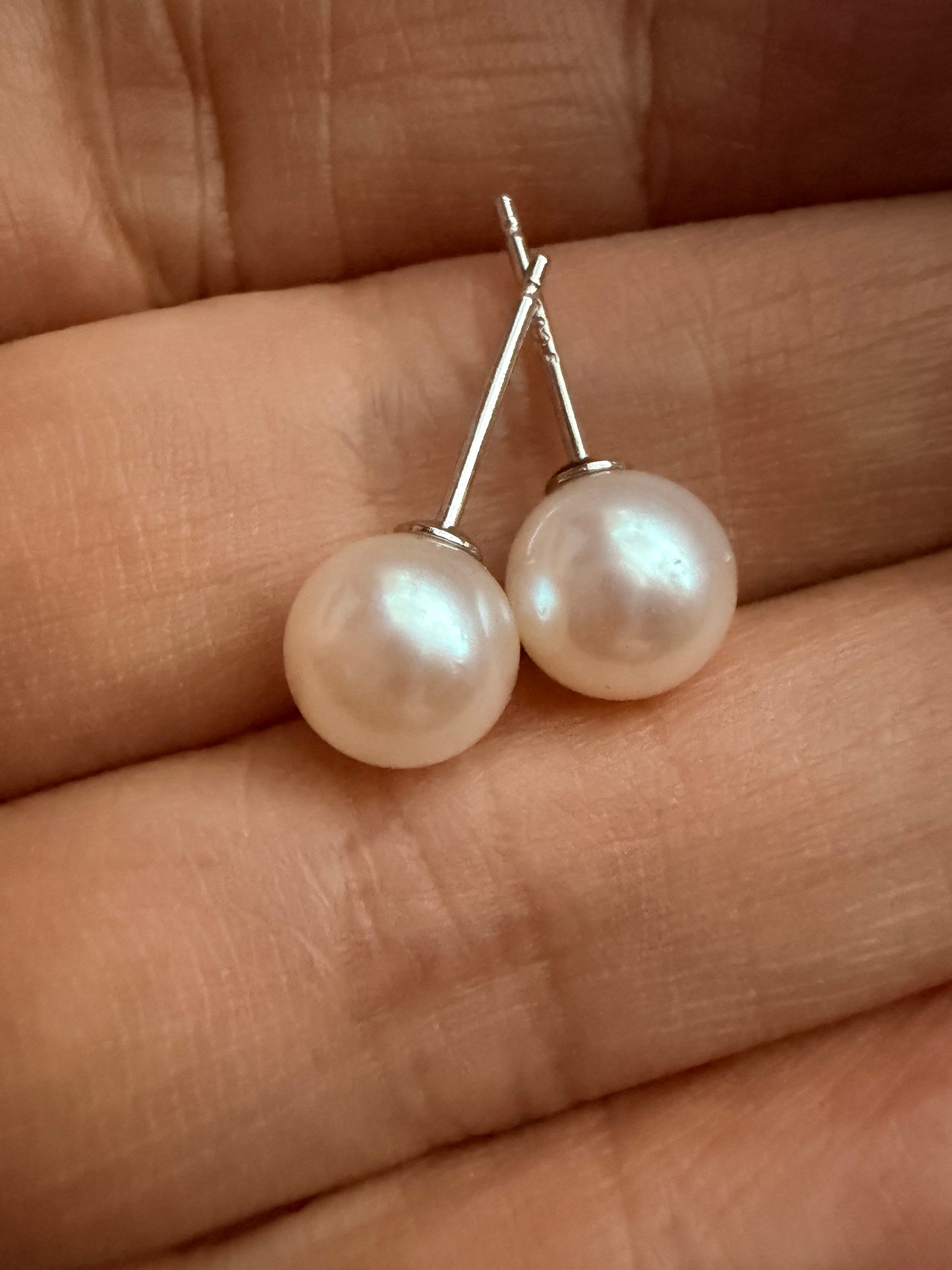 7mm round white saltwater pearl stud earrings with 925 sterling silver posts held in a hand.