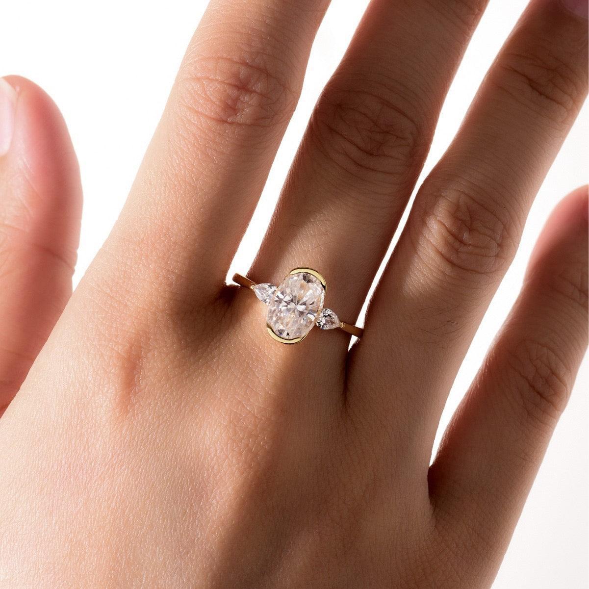 Three-stone moissanite ring with a 7x10mm oval center and pear side stones in a gold half-bezel setting on a finger.