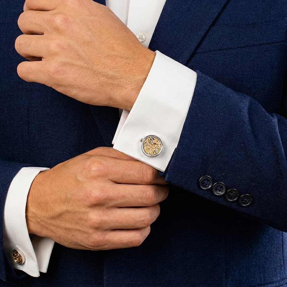 Brass skeleton movement cufflinks with visible gears and ruby bearings, worn on a white shirt cuff under a navy blazer.