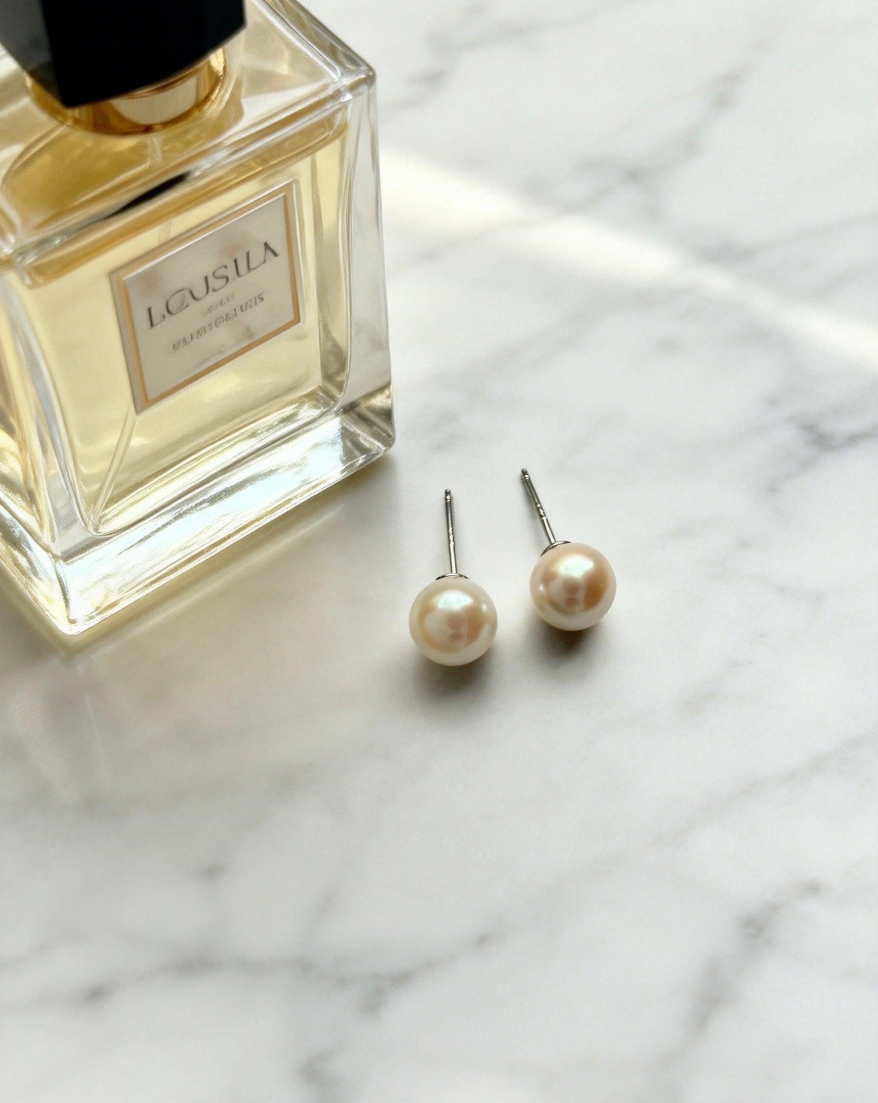 7mm natural saltwater pearl stud earrings with 925 sterling silver posts on a white marble surface next to a perfume bottle.
