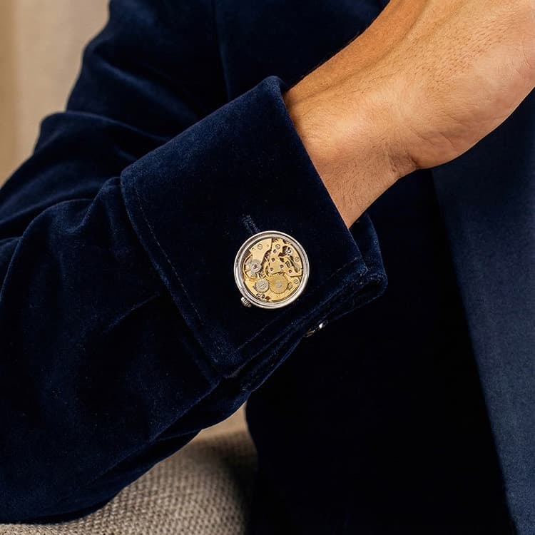Round brass skeleton movement cufflinks with visible gold gears and ruby bearings worn on a navy velvet shirt cuff.