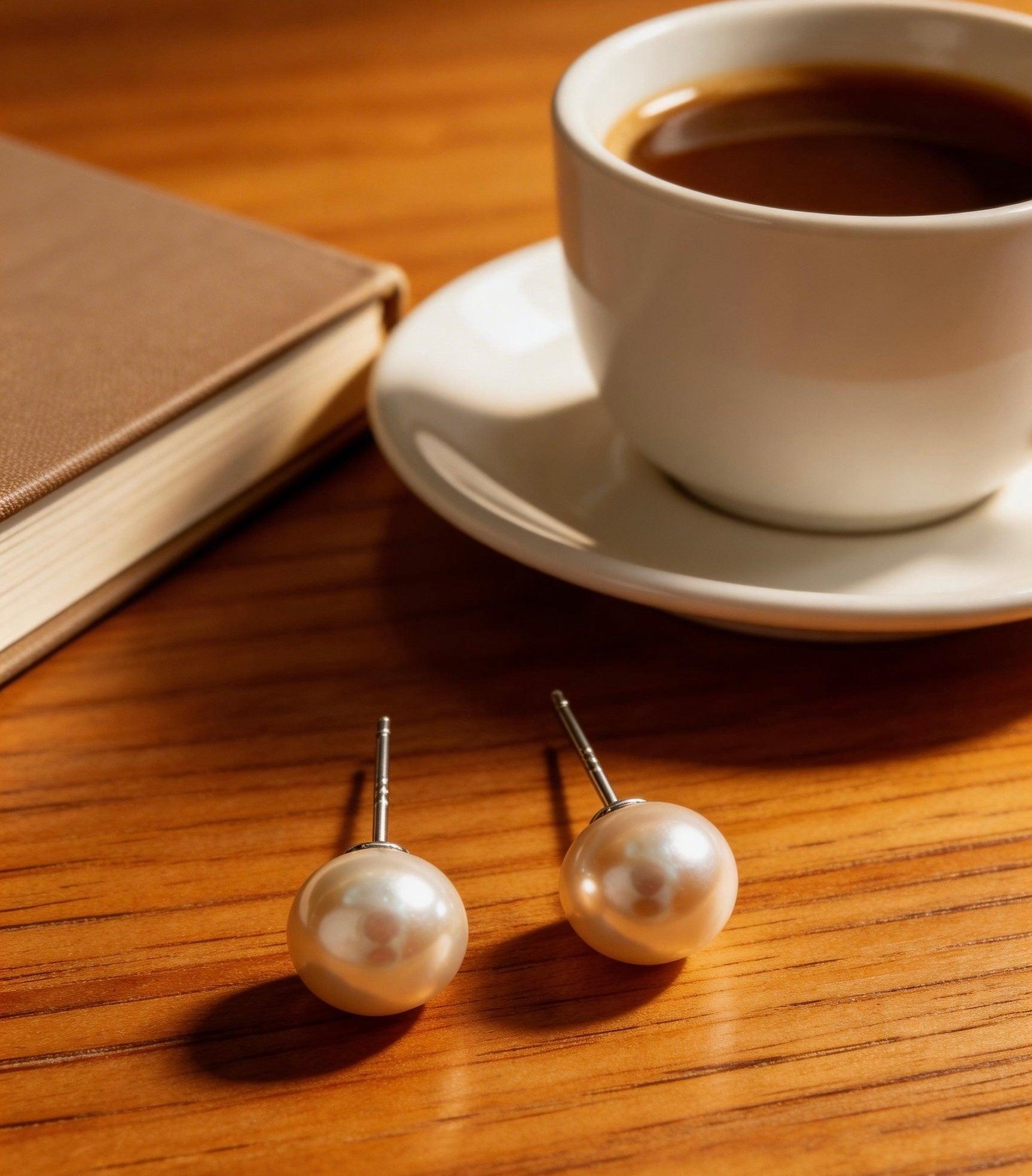 7mm lustrous freshwater pearl stud earrings with sterling silver posts on a wooden table next to a coffee cup and book.